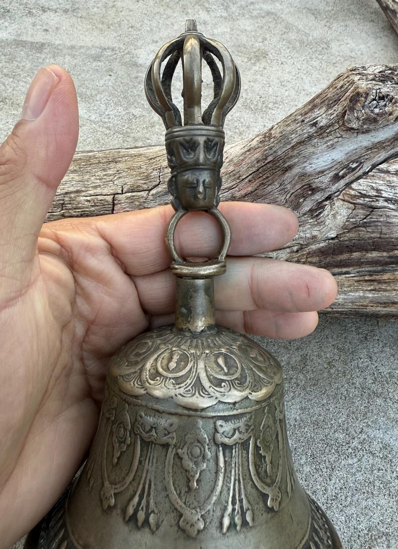 May include: A silver bell with intricate carvings and a crown-shaped handle. The bell has a detailed design of a face on the handle and floral patterns on the body.