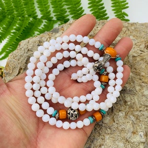 May include: White shell bead mala necklace with orange and turquoise accents. The necklace has a silver charm with intricate details.