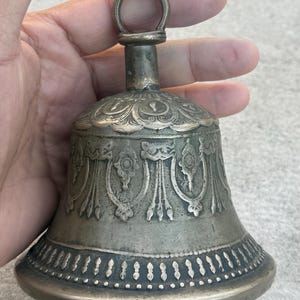 May include: A silver bell with intricate carvings and a decorative handle. The bell has a rounded shape and a wide base.