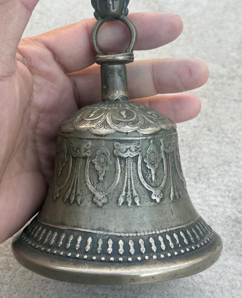 May include: A silver bell with intricate carvings and a decorative handle. The bell has a rounded shape and a wide base.