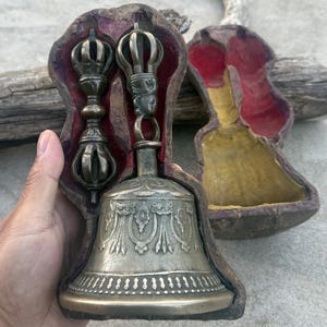 May include: A silver bell with intricate carvings and a red velvet lining inside a wooden box. The bell is accompanied by two silver hand-held ritual objects with ornate designs.