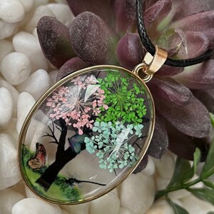Puede incluir: Collar con colgante de forma ovalada y cord&oacute;n negro. El colgante es de vidrio transparente con un borde dorado y contiene flores prensadas y una mariposa.