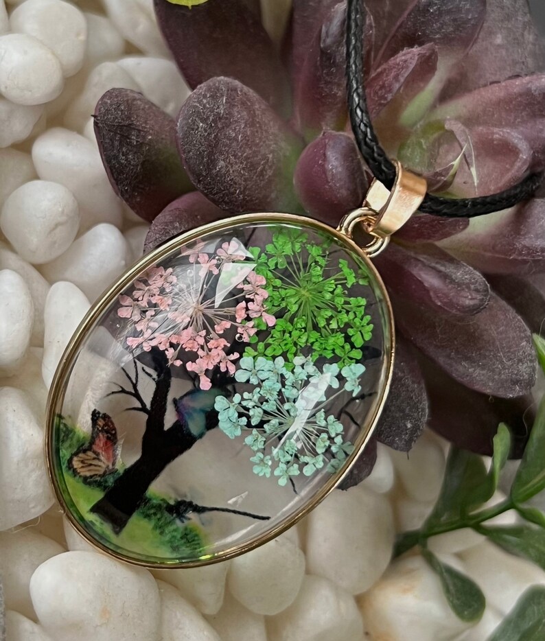 Puede incluir: Collar con colgante de forma ovalada y cord&oacute;n negro. El colgante es de vidrio transparente con un borde dorado y contiene flores prensadas y una mariposa.