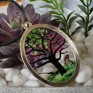 Puede incluir: Un collar con un colgante ovalado de plata con una imagen de resina de un &aacute;rbol con flores rosas y verdes y una mariposa. El colgante est&aacute; en un cord&oacute;n negro.
