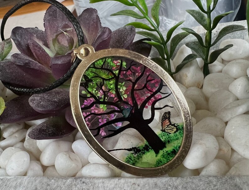 Puede incluir: Un collar con un colgante ovalado de plata con una imagen de resina de un &aacute;rbol con flores rosas y verdes y una mariposa. El colgante est&aacute; en un cord&oacute;n negro.
