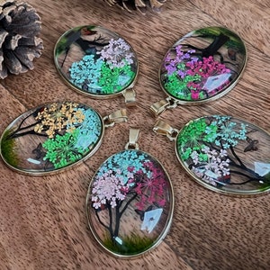Puede incluir: Cinco colgantes de forma ovalada con marcos dorados, cada uno con una disposici&oacute;n diferente de flores prensadas y mariposas en tonos de rosa, verde, amarillo y azul.
