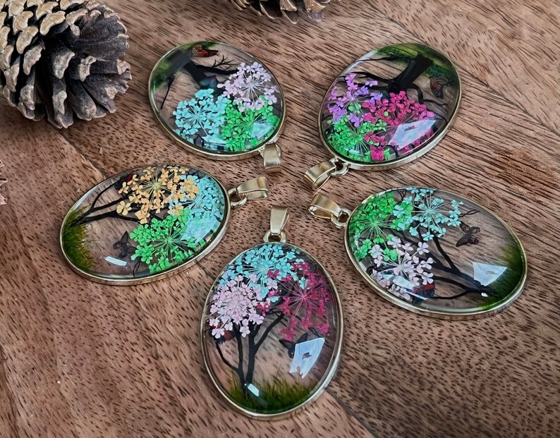 Puede incluir: Cinco colgantes de forma ovalada con marcos dorados, cada uno con una disposici&oacute;n diferente de flores prensadas y mariposas en tonos de rosa, verde, amarillo y azul.