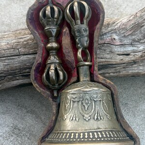 May include: A set of Tibetan Buddhist ritual instruments, a bell and a dorje, in a wooden box with a red velvet lining. The bell is silver with intricate engravings and the dorje is made of brass.