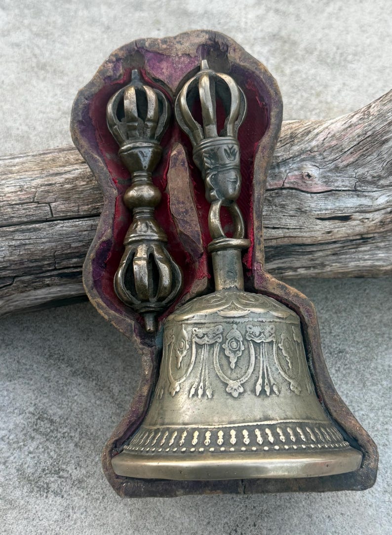 May include: A set of Tibetan Buddhist ritual instruments, a bell and a dorje, in a wooden box with a red velvet lining. The bell is silver with intricate engravings and the dorje is made of brass.