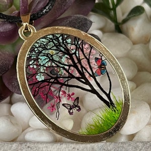 Puede incluir: Un collar con un colgante ovalado de oro con un inserto de resina transparente que presenta una silueta negra de un &aacute;rbol con flores rosas, azules y blancas y dos mariposas.