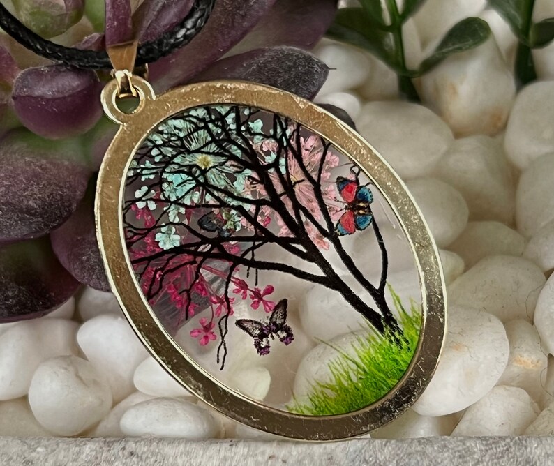 Puede incluir: Un collar con un colgante ovalado de oro con un inserto de resina transparente que presenta una silueta negra de un &aacute;rbol con flores rosas, azules y blancas y dos mariposas.