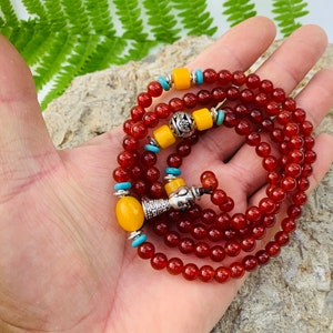 May include: A red carnelian mala prayer bead necklace with yellow and turquoise accents. The necklace has a silver charm and a tassel.