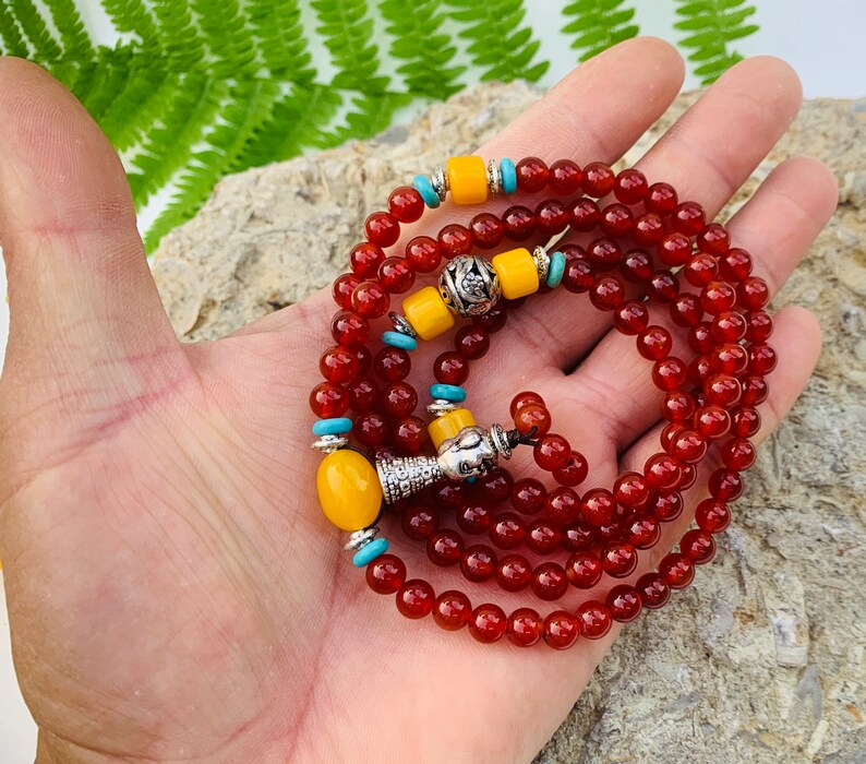May include: A red carnelian mala prayer bead necklace with yellow and turquoise accents. The necklace has a silver charm and a tassel.