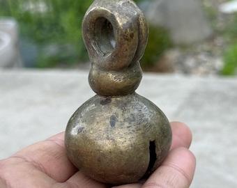 May include: A vintage brass bell with a loop handle. The bell has a rounded body with a slit and a small clapper inside. The brass has a tarnished, aged appearance. The bell is held in a hand.
