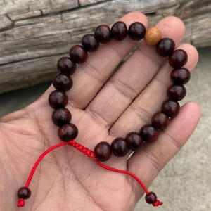 May include: A dark red wooden beaded bracelet with a single light brown wooden bead. The bracelet is tied with a red cord.