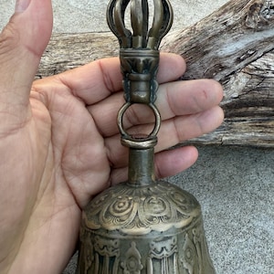 May include: A brass bell with intricate carvings and a decorative finial. The bell has a traditional design and is likely used for religious purposes.