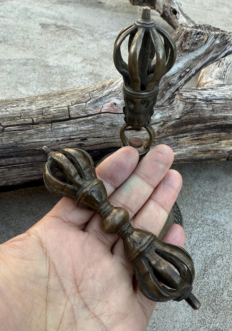 May include: A pair of antique brass Tibetan ritual bells with intricate designs. The bells are connected by a chain and have a handle with a decorative knob.