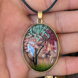 Puede incluir: Un collar con un colgante ovalado de oro y un dije de resina transparente con un dise&ntilde;o de flores prensadas. El dije representa un &aacute;rbol con flores rosas, verdes y rojas y una mariposa.