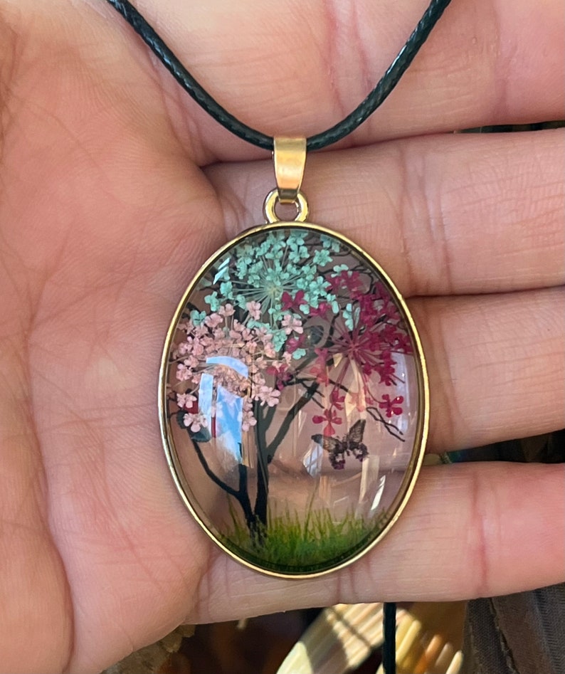 Puede incluir: Un collar con un colgante ovalado de oro y un dije de resina transparente con un dise&ntilde;o de flores prensadas. El dije representa un &aacute;rbol con flores rosas, verdes y rojas y una mariposa.