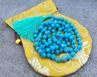 May include: A turquoise blue mala bead necklace with a turquoise tassel, resting in a yellow fabric pouch with an intricate floral design.