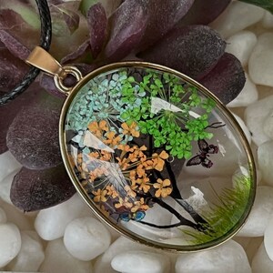 Puede incluir: Un collar con un colgante ovalado de oro y un inserto de resina transparente con flores y mariposas prensadas. Las flores son verdes, naranjas y blancas.