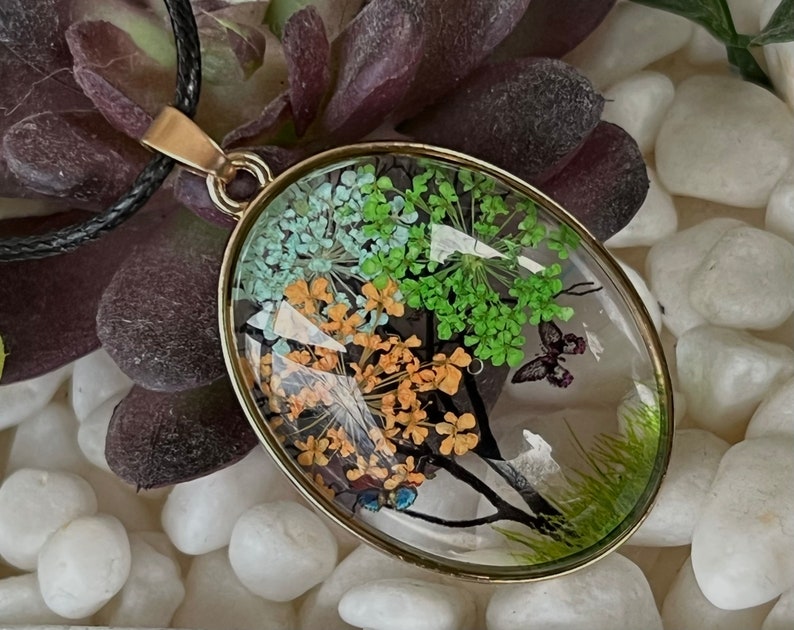 Puede incluir: Un collar con un colgante ovalado de oro y un inserto de resina transparente con flores y mariposas prensadas. Las flores son verdes, naranjas y blancas.