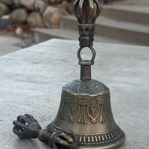 May include: A silver-colored bell with intricate carvings and a matching hand-held clapper. The bell is mounted on a stand with a decorative top.