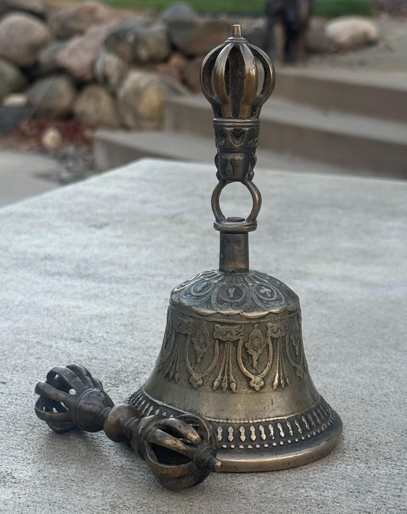 May include: A silver-colored bell with intricate carvings and a matching hand-held clapper. The bell is mounted on a stand with a decorative top.