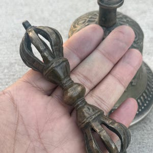 May include: A bronze hand-held bell with a decorative handle. The bell has a detailed design and a small ring at the top. The handle is a long, thin rod with a decorative knob in the middle.