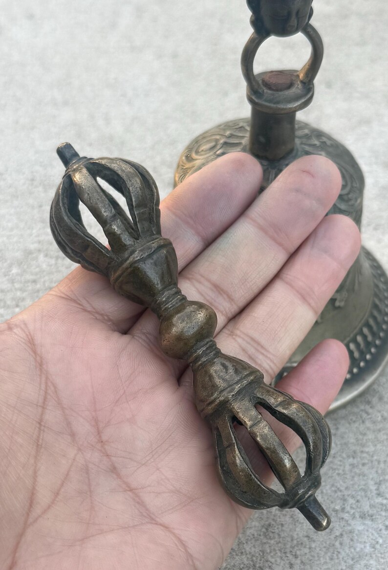 May include: A bronze hand-held bell with a decorative handle. The bell has a detailed design and a small ring at the top. The handle is a long, thin rod with a decorative knob in the middle.