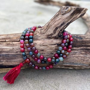 Puede incluir: Un collar de cuentas de varias hebras con cuentas esféricas pulidas en tonos rojo intenso, magenta y verde azulado oscuro. El collar termina con una borla de tela roja vibrante unida a una cuenta roja más grande y lisa en el extremo.