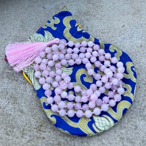 Puede incluir: Un collar mala de cuarzo rosa con un borla rosa, descansando sobre una bolsa de patrón azul y dorado.