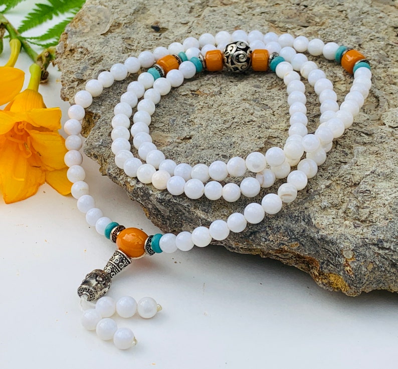 May include: White bead mala necklace with orange and turquoise accents. The necklace has a silver charm at the end.
