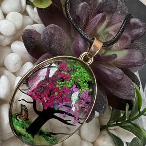 Puede incluir: Un collar con un colgante ovalado de oro en un cord&oacute;n negro. El colgante presenta una c&uacute;pula de resina transparente con una silueta negra de un &aacute;rbol con flores prensadas rosas, moradas y verdes en su interior.