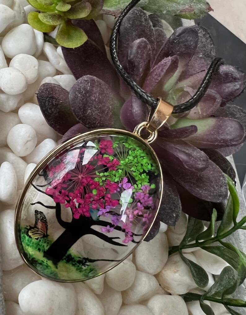 Puede incluir: Un collar con un colgante ovalado de oro en un cord&oacute;n negro. El colgante presenta una c&uacute;pula de resina transparente con una silueta negra de un &aacute;rbol con flores prensadas rosas, moradas y verdes en su interior.
