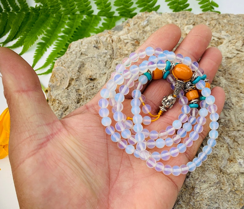 May include: A white opal mala bead necklace with orange and turquoise accents. The necklace is made of small, round beads and has a silver charm at the end.