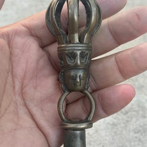 May include: A close-up of the top of a bronze bell with a decorative crown and a face on the handle.