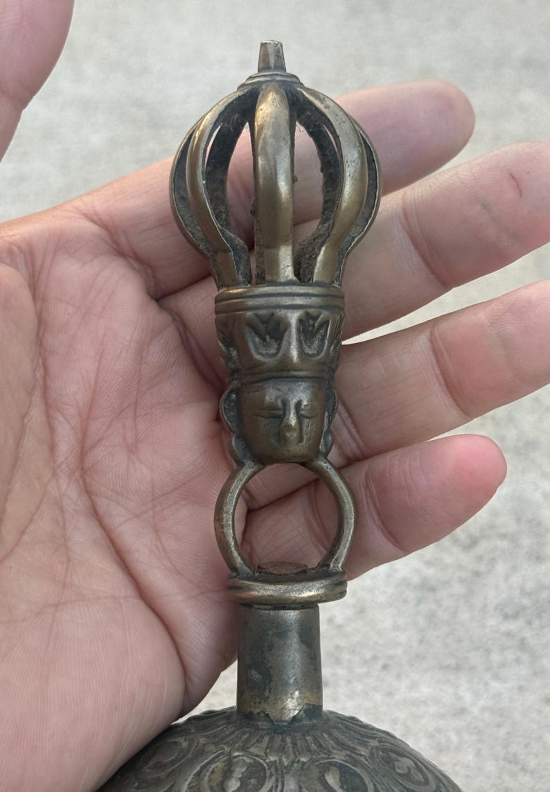 May include: A close-up of the top of a bronze bell with a decorative crown and a face on the handle.