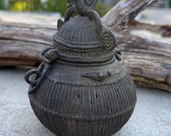 Puede incluir: Una olla oscura de color bronce con tapa y superficie texturizada. La olla presenta un asa en forma de cuerda y una figura decorativa de serpiente en la parte superior. El diseño de la olla incluye líneas verticales y una tapa detallada.