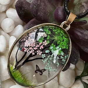 Puede incluir: Un collar con un colgante ovalado de oro y una c&uacute;pula de resina transparente que contiene flores prensadas y una mariposa. Las flores son rosas, verdes y blancas.