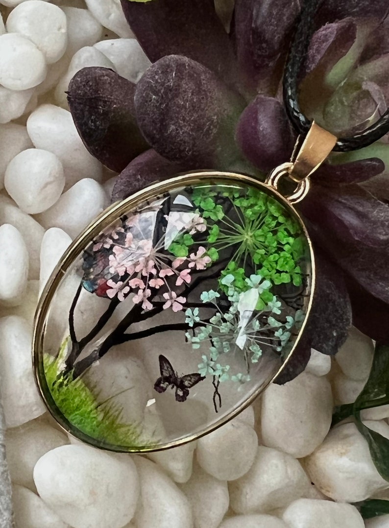 Puede incluir: Un collar con un colgante ovalado de oro y una c&uacute;pula de resina transparente que contiene flores prensadas y una mariposa. Las flores son rosas, verdes y blancas.