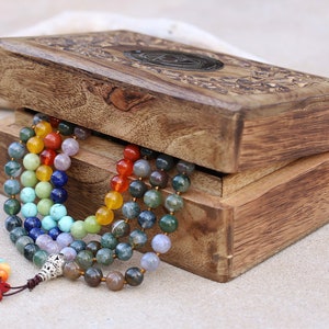 Puede incluir: Una caja de madera con un diseño tallado en la tapa, que contiene un collar de cuentas multicolores. El collar está hecho de varias piedras preciosas, incluyendo amatista, turquesa y cornalina.
