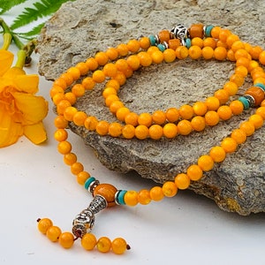 May include: A yellow beaded mala necklace with a silver charm and turquoise accents. The necklace is laid out on a gray rock.