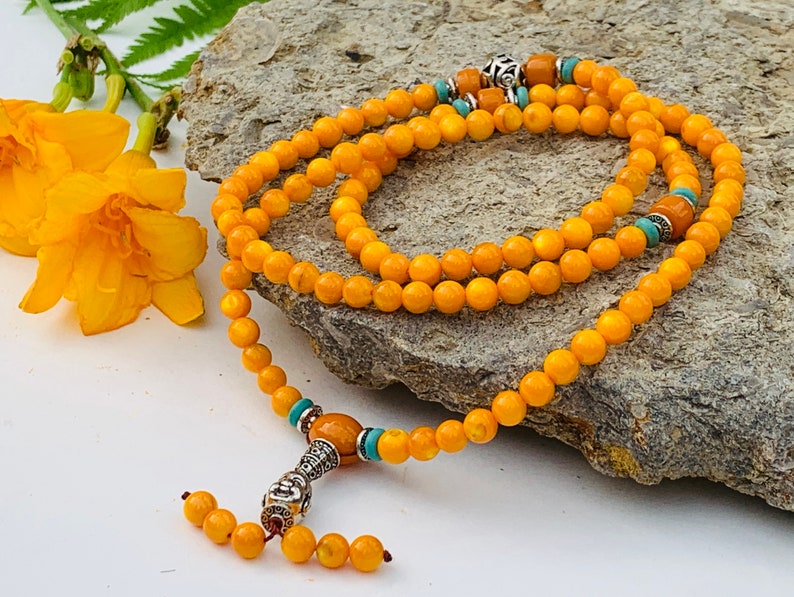 May include: A yellow beaded mala necklace with a silver charm and turquoise accents. The necklace is laid out on a gray rock.