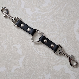 May include: A black strap connector featuring two silver-toned metal swivel trigger snaps at each end. The center of the strap is interrupted by a silver-toned metal heart-shaped ring. Each side of the strap is secured with two silver-toned rivets.