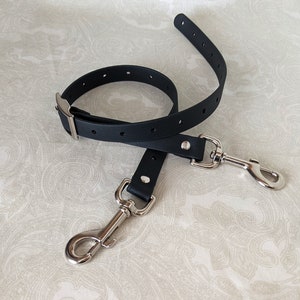 May include: A black leather dog leash with silver metal hardware, including a buckle and a hook. The leash is lying on a white background with a paisley pattern.