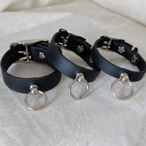 May include: Three black leather collars with silver metal rings and buckles. The collars are on a white fabric surface.
