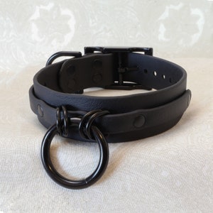 Op de afbeelding: Zwarte leren choker met een grote, zwarte metalen ring eraan bevestigd. De choker heeft een gespsluiting en is verstelbaar.