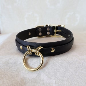 Op de afbeelding: Een zwarte leren choker met een gouden metalen ring en een knoopdetail. De choker heeft een gespsluiting.