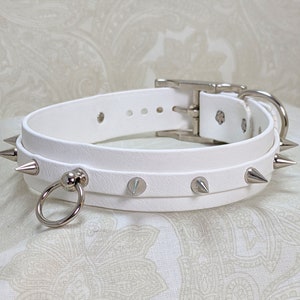 May include: White leather choker with silver spikes and a silver ring. The choker has a buckle closure.